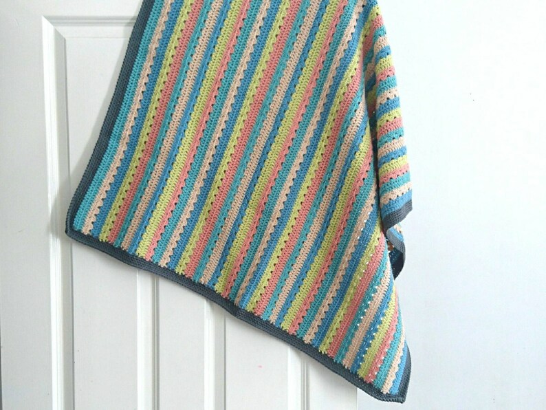 Puede incluir: Una manta de ganchillo multicolor con rayas verticales en tonos pastel de azul, rosa, amarillo y beige. La manta tiene un borde gris y est&aacute; colgada sobre una puerta blanca. El patr&oacute;n de ganchillo presenta un dise&ntilde;o de puntada abierta y texturizada.
