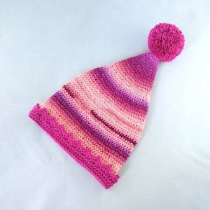 May include: A handmade, pointed crochet hat in shades of pink and purple, topped with a large, matching pom-pom. The hat features a scalloped edge in a darker pink, adding a decorative touch. The gradient color scheme creates a visually appealing effect.