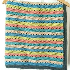 Puede incluir: Una colorida manta de ganchillo hecha a mano con rayas horizontales en tonos de azul, rosa, amarillo y beige. La manta tiene un borde festoneado y se muestra en una percha de madera. La manta mide aproximadamente 100 cm de ancho y 127 cm de largo.