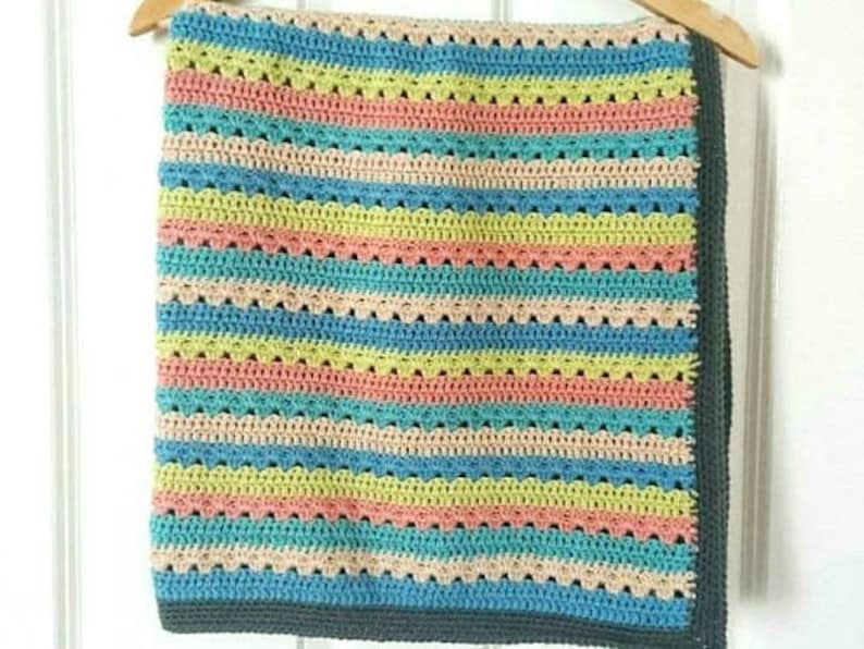 Puede incluir: Una colorida manta de ganchillo hecha a mano con rayas horizontales en tonos de azul, rosa, amarillo y beige. La manta tiene un borde festoneado y se muestra en una percha de madera. La manta mide aproximadamente 100 cm de ancho y 127 cm de largo.
