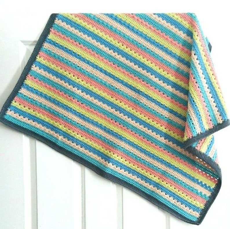 Puede incluir: Una manta de ganchillo hecha a mano con un patr&oacute;n de rayas. La manta presenta filas de colores pastel, incluyendo azul, amarillo, rosa y crema, con un borde gris oscuro. La manta est&aacute; doblada, mostrando su textura y dise&ntilde;o.