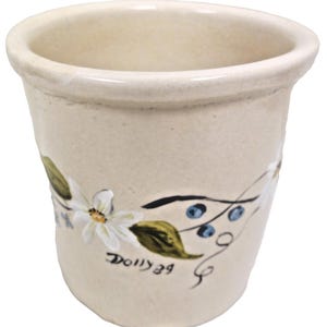 May include: A cream-colored ceramic vase with a floral design. The vase features white flowers, green leaves, and blue berries. The text "Dolly 39" is hand-painted on the side. The vase has a slightly flared rim and a smooth, unglazed finish.