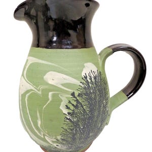 May include: Handmade ceramic pitcher with a black rim and handle. The body is green with white marbling and a black tree-like design. The pitcher has a spout for easy pouring and a sturdy handle.