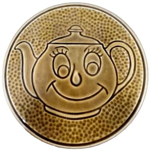 May include: A round, ceramic trivet with a smiling teapot design. The teapot is light brown with a dark brown outline and features large eyes and a smiling mouth. The background is a textured, dotted pattern in a similar shade of brown.