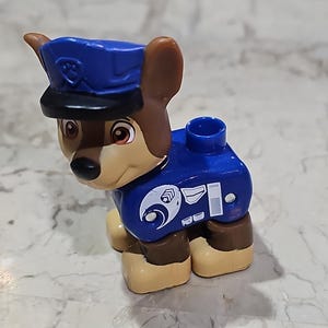 May include: A blue and brown plastic toy dog figure wearing a blue police hat with a paw print on it. The toy has a white paw print on its chest and a white star on its side.