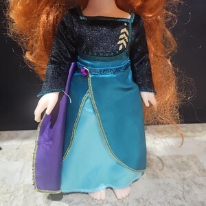 Puede incluir: Una mu&ntilde;eca con un vestido azul y verde con una capa morada. El vestido tiene ribetes dorados y un corpi&ntilde;o negro con bordados de hojas doradas.