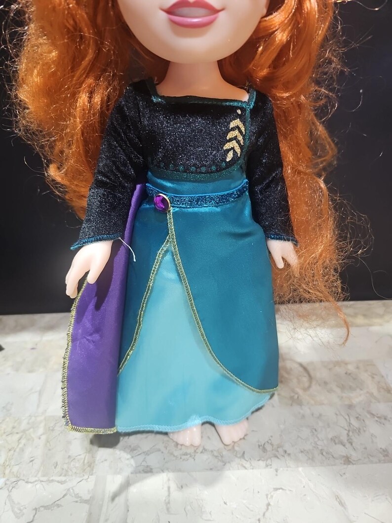 Puede incluir: Una mu&ntilde;eca con un vestido azul y verde con una capa morada. El vestido tiene ribetes dorados y un corpi&ntilde;o negro con bordados de hojas doradas.