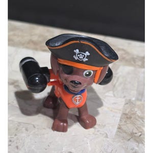 May include: A brown dog figurine dressed as a pirate, wearing a black pirate hat with a white skull and paw print emblem, an orange vest with a silver anchor badge, and a black eye patch. The figure has black cylindrical attachments on its sides.