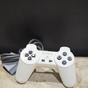 Könnte beinhalten: Ein weißer Sony PlayStation-Controller mit einem grauen Kabel. Der Controller hat einen schwarzen Richtungspfeil, vier schwarze Fronttasten und zwei schwarze Tasten mit der Aufschrift "Select" und "Start".