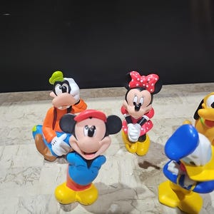 May include: A set of five colorful plastic Disney character figurines. The characters include Mickey Mouse, Minnie Mouse, Goofy, Donald Duck, and Pluto. The figurines are all in a standing position and have a cartoonish style.
