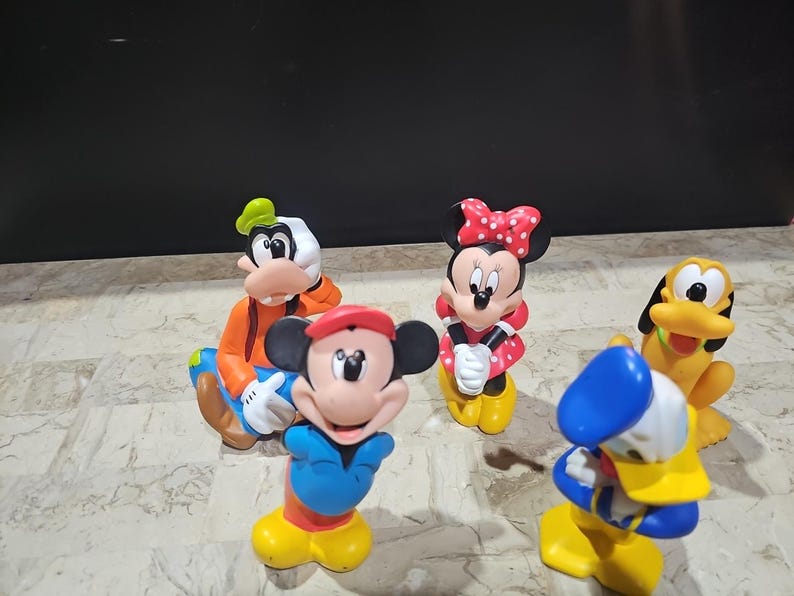 May include: A set of five colorful plastic Disney character figurines. The characters include Mickey Mouse, Minnie Mouse, Goofy, Donald Duck, and Pluto. The figurines are all in a standing position and have a cartoonish style.