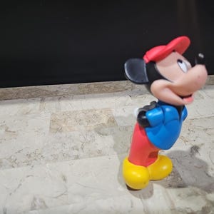 May include: A red, blue, and yellow plastic toy figurine of Mickey Mouse wearing a red hat and a blue jacket.