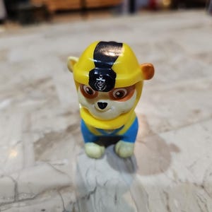 May include: A yellow and blue toy dog wearing a black and yellow construction helmet. The dog has brown ears and a black nose.