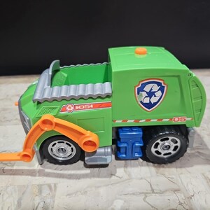 May include: Green toy garbage truck with an orange arm, a blue recycling bin, and the number 05 on the side. The truck has a paw print and the words "K054" on the side.