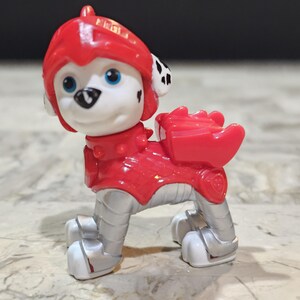 May include: A red and silver toy dog wearing a helmet and armour. The dog has black spots and white fur. The toy is part of a popular children's cartoon.