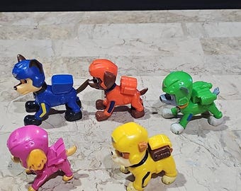 May include: Five plastic toy figurines of dogs wearing different colored helmets and uniforms. The dogs are in various poses, including standing, sitting, and running. The figurines are part of a popular children's television show.