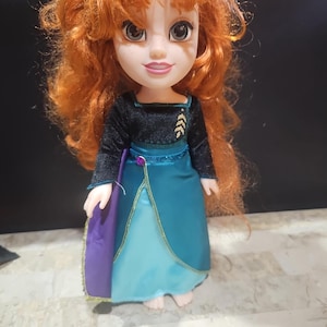 Peut inclure: Une poupée aux cheveux roux portant une robe bleue et noire avec des accents dorés. La poupée a les yeux bruns et un visage souriant.