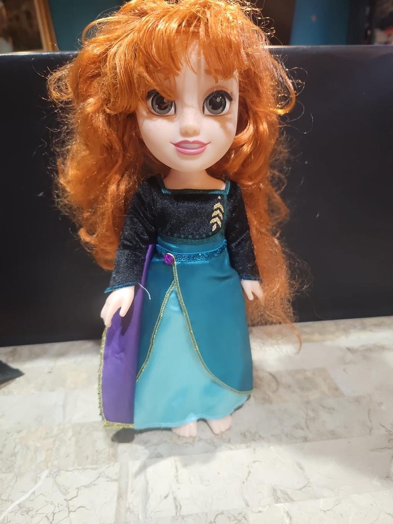 Puede incluir: Una mu&ntilde;eca de pelo rojo con un vestido azul y negro con detalles dorados. La mu&ntilde;eca tiene ojos marrones y una cara sonriente.