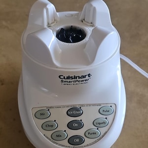 Puede incluir: Una licuadora electrónica blanca Cuisinart SmartPower de 7 velocidades. El panel de control tiene botones para Stir, Chop, Mix, Ice Crush, On, Off, Pulse, Liquefy y Purée. Una advertencia está impresa encima de los botones.