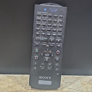 Puede incluir: Un mando a distancia negro para DVD de PlayStation 2 con botones para mezclar audio, programar ángulo, repetir subtítulos, lento, borrar, hora, escanear, anterior, siguiente, A+B, reproducir, pausa, detener, mostrar, título, menú DVD, volver, entrar, triángulo, cuadrado, círculo, X, L1, L2, L3, R1, R2, R3, seleccionar, iniciar y un logotipo de Sony con el texto "DVD/PLAYSTATION".