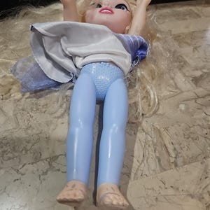 May include: A blonde doll with blue leggings and a white and blue dress. The doll is lying on its back with its arms raised above its head.