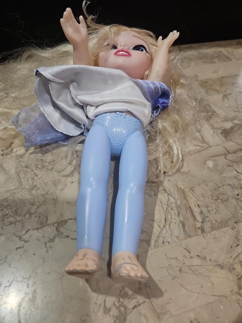 May include: A blonde doll with blue leggings and a white and blue dress. The doll is lying on its back with its arms raised above its head.