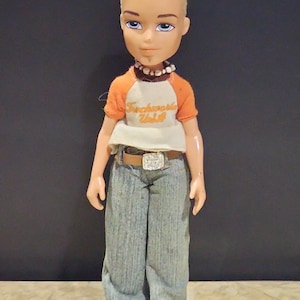 Könnte beinhalten: Eine Bratz-Puppe mit kurzen, gestylten blonden Haaren und blauen Augen. Die Puppe trägt ein orangefarbenes und weißes kurzärmeliges T-Shirt mit dem Text "Suchworld Wild", eine blaue Jeans, einen braunen Gürtel mit silberner Schnalle, eine Perlenkette und blaue Turnschuhe.