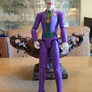 Peut inclure: Figurine du Joker en costume violet, gilet vert et chemise orange. La figurine a les cheveux verts, la peau blanche et un sourire rouge. Des chaussures noires complètent le look. La figurine est sur une table.