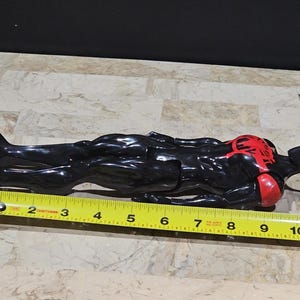 May include: A black and red Spider-Man action figure, approximately 11 inches long, lies on a surface. The figure has a red spider emblem on the chest and red accents on the arms. A yellow measuring tape is placed next to the figure.