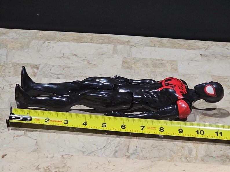May include: A black and red Spider-Man action figure, approximately 11 inches long, lies on a surface. The figure has a red spider emblem on the chest and red accents on the arms. A yellow measuring tape is placed next to the figure.