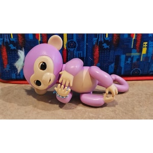 May include: A purple toy monkey with a light tan belly and articulated limbs. The monkey has large black eyes, a white nose, and a tuft of blue hair. A blue bracelet adorns its wrist. The toy is lying on a light brown surface.
