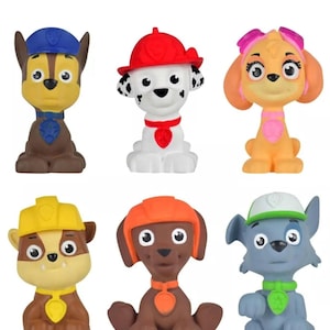 May include: Six colourful Paw Patrol character figurines. Each figure is a different colour, including blue, red, orange, yellow, and pink. The figurines are in a sitting position and have large eyes and distinct features.