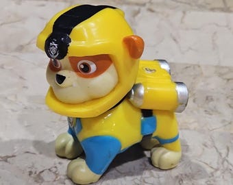 May include: A yellow and blue toy dog figure with a black stripe on its helmet. The toy has orange accents around the eyes and ears, and a silver pack on its back. The figure is standing on a marble-like surface.
