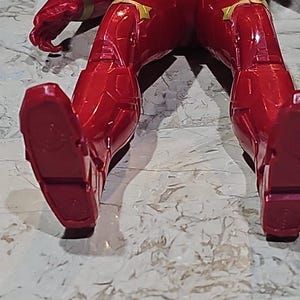 Pu&ograve; includere: Figurina d'azione rossa e dorata con un costume rosso e nero. La figura &egrave; sdraiata sulla schiena con le gambe in aria. I piedi della figura sono visibili.