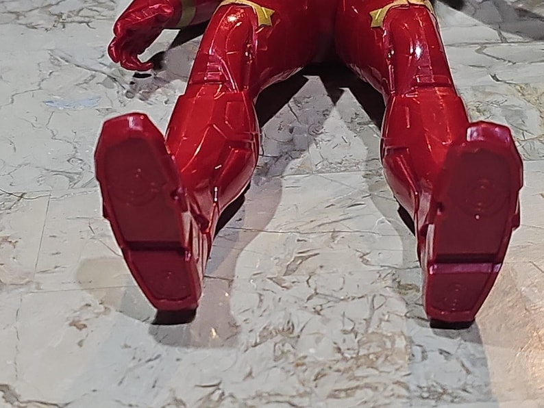Pu&ograve; includere: Figurina d'azione rossa e dorata con un costume rosso e nero. La figura &egrave; sdraiata sulla schiena con le gambe in aria. I piedi della figura sono visibili.