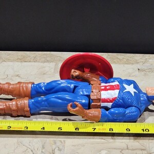 May include: A blue and red action figure of Captain America lying on its back with a red and white shield behind it. The figure is approximately 12 inches long.