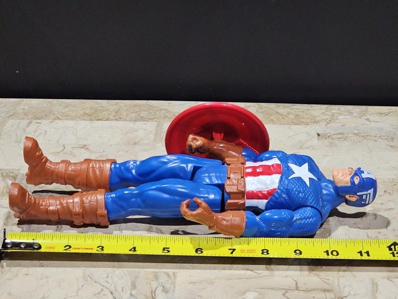 May include: A blue and red action figure of Captain America lying on its back with a red and white shield behind it. The figure is approximately 12 inches long.
