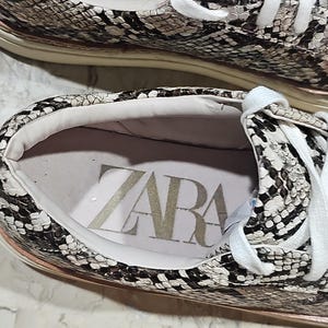 May include: Close-up of a pair of sneakers with a snakeskin pattern in shades of brown, gray, and white. The interior of the shoe is visible, with the brand name "ZARA" printed in large letters. White laces and a light-colored sole complete the design.