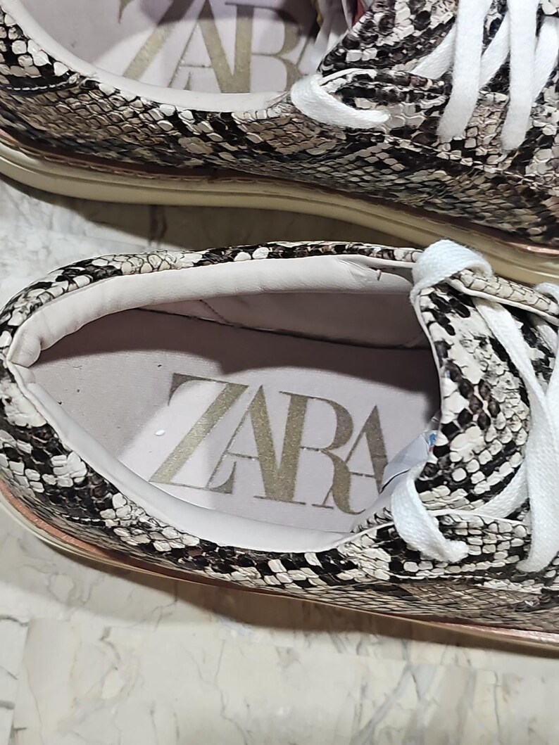 May include: Close-up of a pair of sneakers with a snakeskin pattern in shades of brown, gray, and white. The interior of the shoe is visible, with the brand name "ZARA" printed in large letters. White laces and a light-colored sole complete the design.