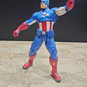 May include: A blue and red action figure of Captain America in a fighting pose. The figure has a red, white, and blue costume with a star on the chest. The figure is holding its right arm out in front of it, with its fist clenched.