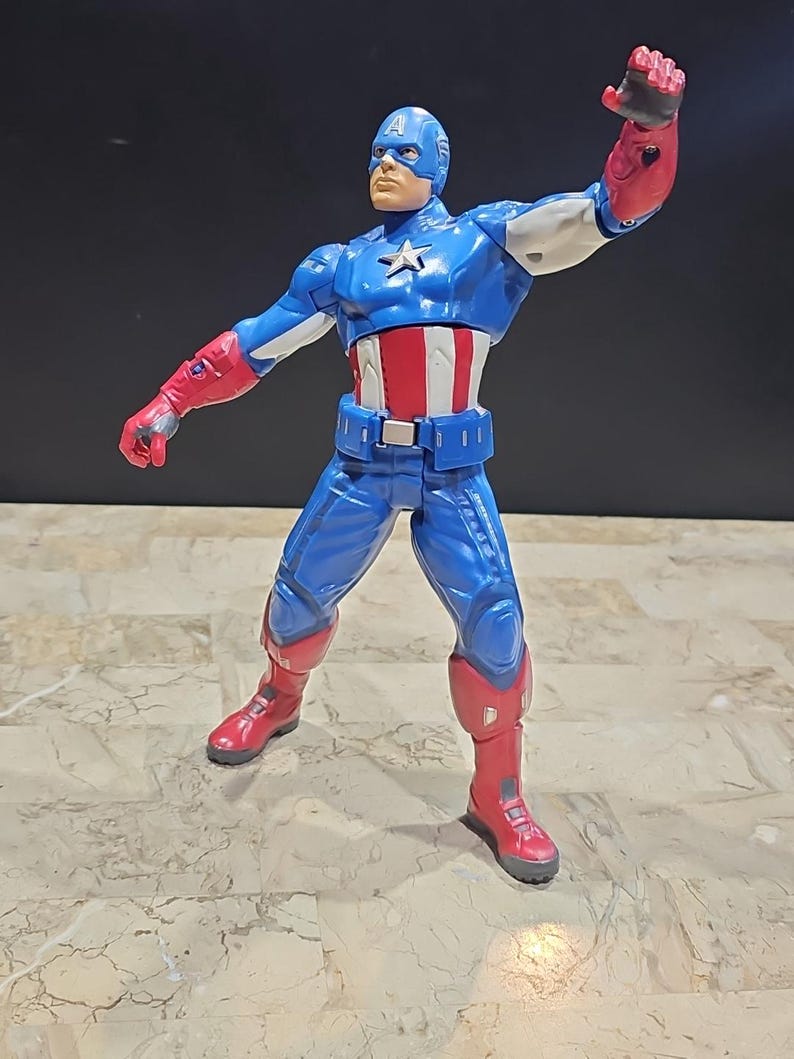 May include: A blue and red action figure of Captain America in a fighting pose. The figure has a red, white, and blue costume with a star on the chest. The figure is holding its right arm out in front of it, with its fist clenched.