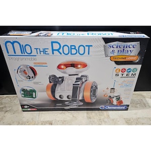 Op de afbeelding: Een doos met Mio the Robot speelgoed, in wit en oranje. De doos toont de tekst "Mio the Robot" en "science & play". De robot heeft oranje wielen en een oplichtend oranje hoofd. De doos toont ook schema's van de robotonderdelen.