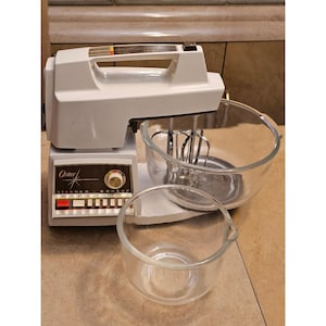 May include: A vintage Oster Kitchen Center stand mixer in white with a clear glass mixing bowl and a smaller clear glass bowl. The mixer has a control panel with the Oster logo and buttons. The mixer is on a beige countertop.