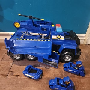 May include: A large blue toy truck with a helicopter on top and several small blue motorcycles. The truck has a door open, revealing the interior. The toy is designed for children.