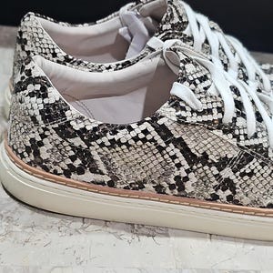 May include: Close-up of a pair of sneakers with a snakeskin pattern in black, white, and gray. The shoes have white laces and a cream-colored sole with a thin pink stripe.