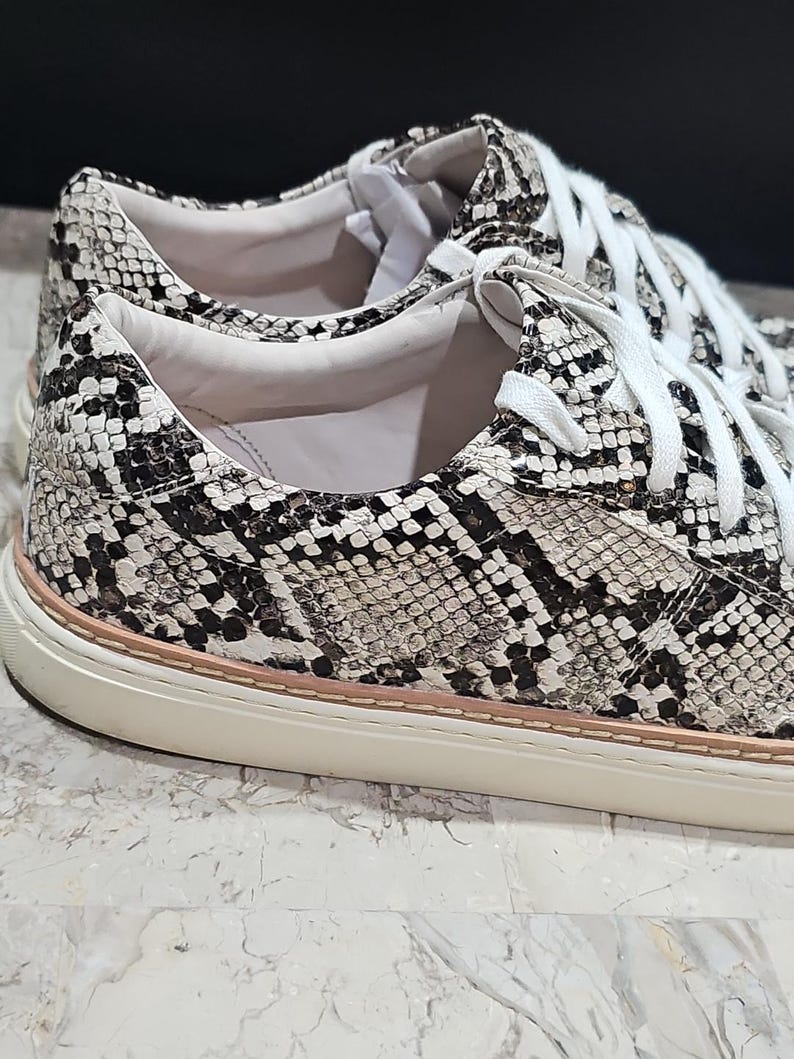 May include: Close-up of a pair of sneakers with a snakeskin pattern in black, white, and gray. The shoes have white laces and a cream-colored sole with a thin pink stripe.