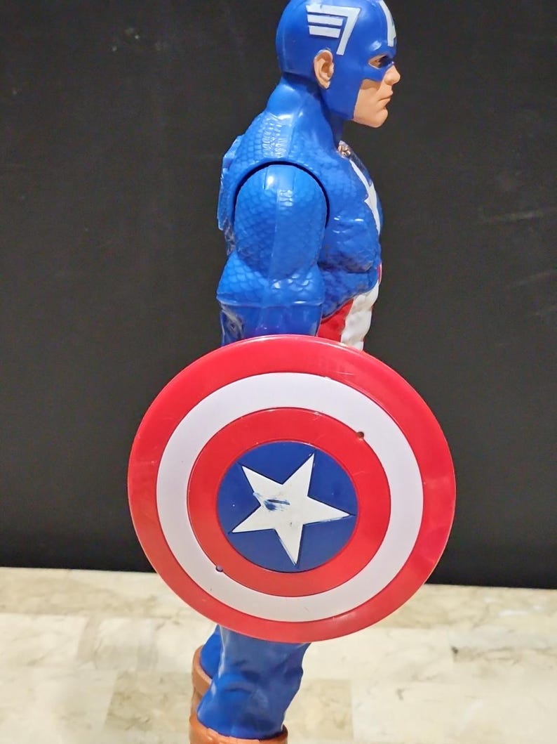 May include: A blue and red plastic action figure of Captain America holding his red, white, and blue shield. The figure has a red, white, and blue costume with a star on the chest. The shield has a white star in the center.