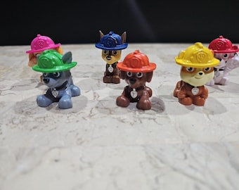 May include: A collection of colorful Paw Patrol figurines. Each puppy is wearing a different colored hat, including pink, green, blue, orange, and yellow. The figurines are set on a light-colored, textured surface.