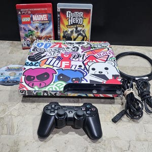 Puede incluir: Una consola PlayStation 3 negra con una parte superior cubierta de pegatinas, un mando negro y discos de juego. Los juegos incluyen "Lego Marvel Super Heroes" y "Guitar Hero World Tour". También se ven cables y un cable de alimentación.