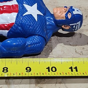 May include: A blue and red Captain America action figure with a white star on his chest. The figure is lying on its side and is approximately 8 inches long. A yellow measuring tape is in the foreground with the numbers 8, 9, 10, 11, and 12 visible.